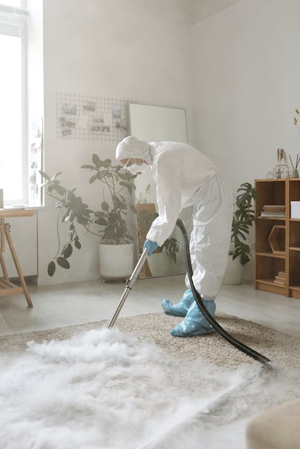 A professional cleaner dressed in a white protective suit, gloves, and shoe covers performs deep cleaning on a beige carpet in a bright, minimalist living room. The room features white walls, a large 