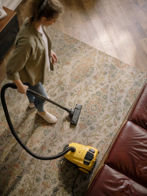 A professional cleaner dressed in a white protective suit, gloves, and shoe covers performs deep cleaning on a beige carpet in a bright, minimalist living room. The room features white walls, a large 